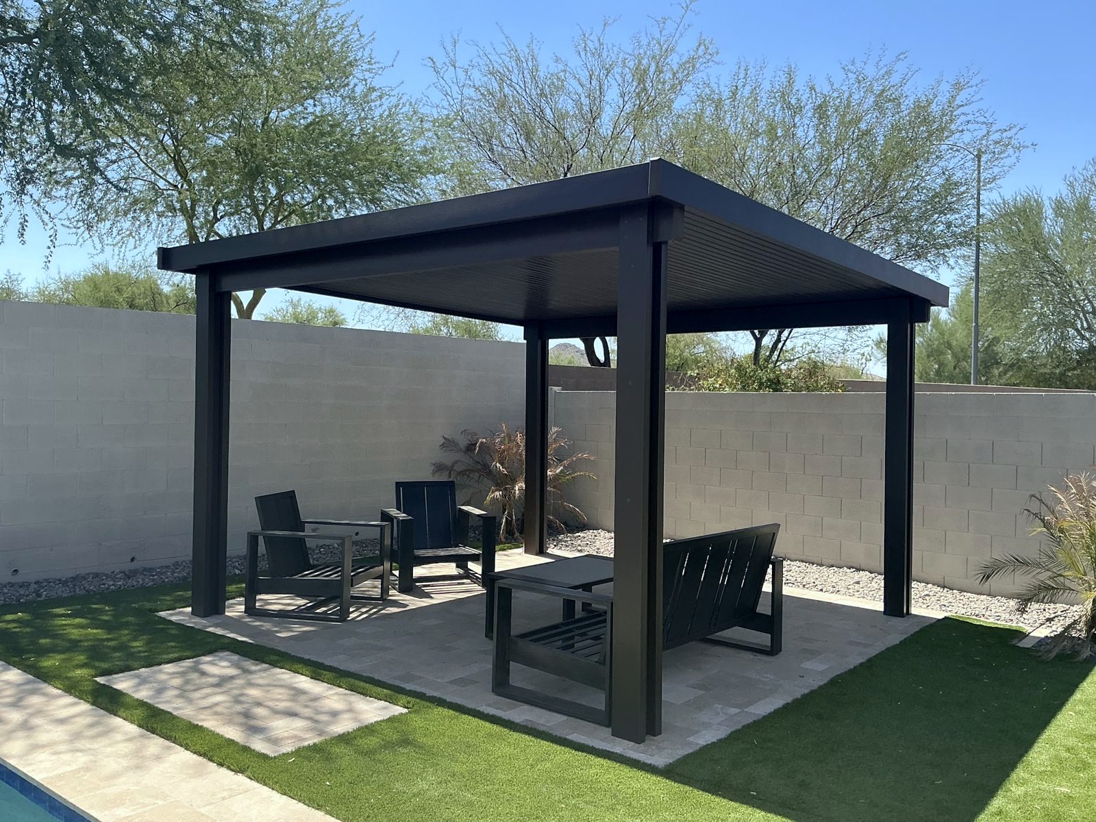 Black aluminum pergola over a travertine lounge pad by the pool